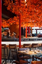 A restaurant setting with a vibrant orange autumn-themed decor featuring numerous artificial leaves and lights adorning branches above the dining area. There are barstools and a set table with wine glasses and napkins ready for diners. Two individuals behind the bar, one showing peace signs, suggest a lively and welcoming atmosphere.