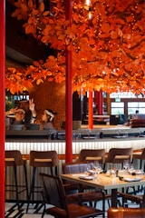 A restaurant setting with a vibrant orange autumn-themed decor featuring numerous artificial leaves and lights adorning branches above the dining area. There are barstools and a set table with wine glasses and napkins ready for diners. Two individuals behind the bar, one showing peace signs, suggest a lively and welcoming atmosphere.