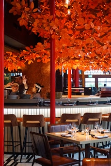 A restaurant setting with a vibrant orange autumn-themed decor featuring numerous artificial leaves and lights adorning branches above the dining area. There are barstools and a set table with wine glasses and napkins ready for diners. Two individuals behind the bar, one showing peace signs, suggest a lively and welcoming atmosphere.
