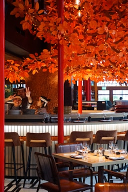 A restaurant setting with a vibrant orange autumn-themed decor featuring numerous artificial leaves and lights adorning branches above the dining area. There are barstools and a set table with wine glasses and napkins ready for diners. Two individuals behind the bar, one showing peace signs, suggest a lively and welcoming atmosphere.