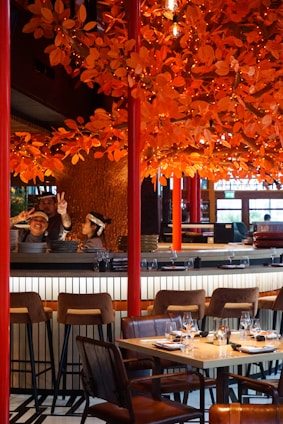A restaurant setting with a vibrant orange autumn-themed decor featuring numerous artificial leaves and lights adorning branches above the dining area. There are barstools and a set table with wine glasses and napkins ready for diners. Two individuals behind the bar, one showing peace signs, suggest a lively and welcoming atmosphere.