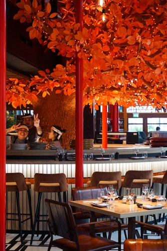 A restaurant setting with a vibrant orange autumn-themed decor featuring numerous artificial leaves and lights adorning branches above the dining area. There are barstools and a set table with wine glasses and napkins ready for diners. Two individuals behind the bar, one showing peace signs, suggest a lively and welcoming atmosphere.