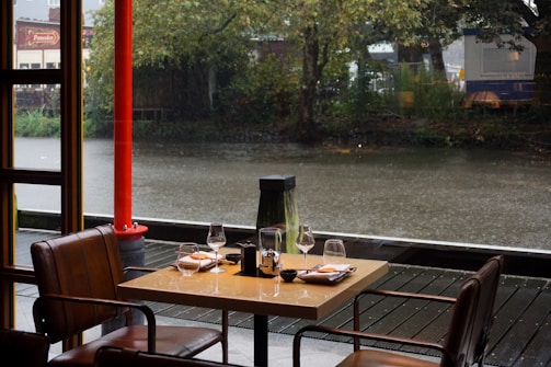 A cozy indoor dining area at a restaurant overlooking a tranquil river. There are two modern leather chairs and a square table set with wine glasses, plates, and folded napkins. It is raining outside, with droplets visible on the water surface. Tall trees line the riverbank, adding a natural backdrop to the scene. A bright red pillar adds a pop of color.