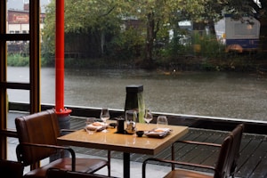 A cozy indoor dining area at a restaurant overlooking a tranquil river. There are two modern leather chairs and a square table set with wine glasses, plates, and folded napkins. It is raining outside, with droplets visible on the water surface. Tall trees line the riverbank, adding a natural backdrop to the scene. A bright red pillar adds a pop of color.