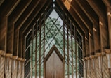 An intricate wooden structure with large geometric windows framing a lush green forest view. The architecture features pointed arches and a symmetrical design, creating a serene and harmonious atmosphere.