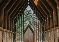 An intricate wooden structure with large geometric windows framing a lush green forest view. The architecture features pointed arches and a symmetrical design, creating a serene and harmonious atmosphere.
