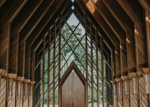 An intricate wooden structure with large geometric windows framing a lush green forest view. The architecture features pointed arches and a symmetrical design, creating a serene and harmonious atmosphere.