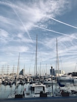 A bustling marina filled with various sailboats and yachts, moored side by side. Tall masts rise against a backdrop of a blue sky with scattered clouds. In the distance, a mix of modern buildings and industrial structures frames the harbor scene.