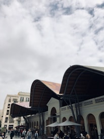 A contemporary market structure with a wave-like roof design covers a plaza where people are walking and gathering. The building features arches and large windows, giving it a blend of modern and classical architectural styles. Overcast skies lend a subdued atmosphere to the scene.
