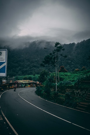 A winding road curves through a lush, green landscape, surrounded by dense forest and mist-covered mountains. A small street market with colorful stalls lines one side of the road. The sky is overcast, creating a moody atmosphere.
