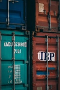 Two shipping containers, one with a teal color and identification numbers, and the other in faded red with a logo. Both containers have metallic structures and locks, indicating industrial use.