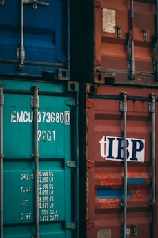 Two shipping containers, one with a teal color and identification numbers, and the other in faded red with a logo. Both containers have metallic structures and locks, indicating industrial use.
