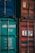 Two shipping containers, one with a teal color and identification numbers, and the other in faded red with a logo. Both containers have metallic structures and locks, indicating industrial use.