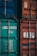 Two shipping containers, one with a teal color and identification numbers, and the other in faded red with a logo. Both containers have metallic structures and locks, indicating industrial use.