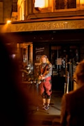 A street musician performs in front of a warmly lit French bakery titled 'Boulangerie Saint Michel'. The artist is dressed in colorful clothing, playing a guitar and singing into a microphone.