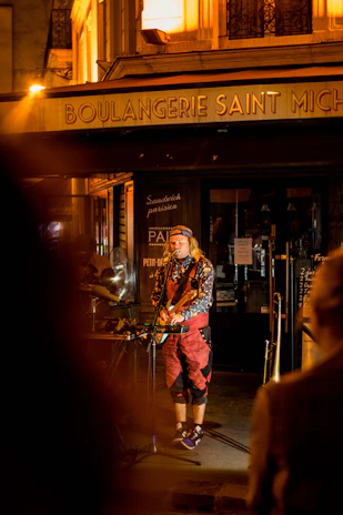 A street musician performs in front of a warmly lit French bakery titled 'Boulangerie Saint Michel'. The artist is dressed in colorful clothing, playing a guitar and singing into a microphone.
