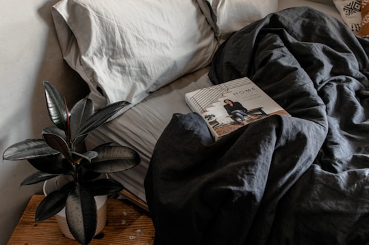 A cozy and inviting bedroom scene features a neatly made bed with soft gray pillows and a dark, rumpled duvet. A glossy-leafed plant in a white pot rests on a wooden bedside table. On the bed lies a magazine titled 'Home' with a photograph of an interior setting on the cover.