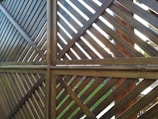 A wooden fence structure made from diagonal slats forms a pattern of intersecting lines. The lighting creates a contrast between the lighter and darker shades of the wood. Some greenery and parts of a garden can be seen in the background through the slats.