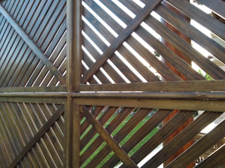 A wooden fence structure made from diagonal slats forms a pattern of intersecting lines. The lighting creates a contrast between the lighter and darker shades of the wood. Some greenery and parts of a garden can be seen in the background through the slats.