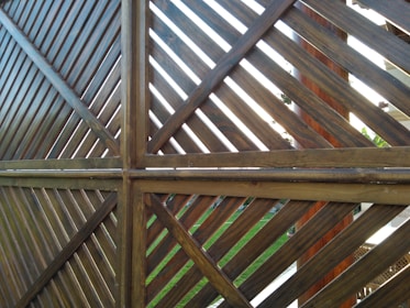 A wooden fence structure made from diagonal slats forms a pattern of intersecting lines. The lighting creates a contrast between the lighter and darker shades of the wood. Some greenery and parts of a garden can be seen in the background through the slats.