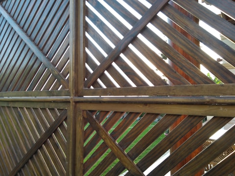 A wooden fence structure made from diagonal slats forms a pattern of intersecting lines. The lighting creates a contrast between the lighter and darker shades of the wood. Some greenery and parts of a garden can be seen in the background through the slats.