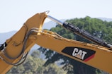 A close-up view of a construction vehicle's hydraulic arm, painted in yellow with the CAT logo prominently visible. In the background, there are blurred trees and mountains, suggesting an outdoor environment.