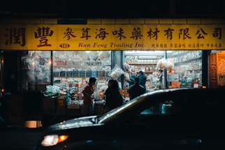 A brightly lit storefront at night with a yellow sign featuring both Chinese characters and English text. Several people are standing in front of the shop, possibly engaged in conversation or purchasing goods. The shop appears to sell various packaged and bulk goods, with shelves inside visible through the window. A car is partially visible in the foreground, adding to the urban, busy atmosphere.