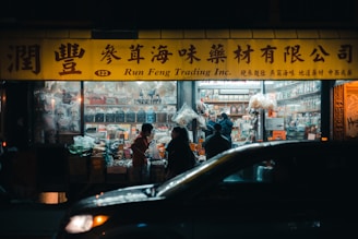 A brightly lit storefront at night with a yellow sign featuring both Chinese characters and English text. Several people are standing in front of the shop, possibly engaged in conversation or purchasing goods. The shop appears to sell various packaged and bulk goods, with shelves inside visible through the window. A car is partially visible in the foreground, adding to the urban, busy atmosphere.