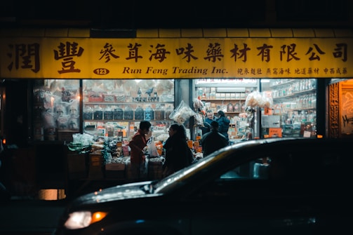 A brightly lit storefront at night with a yellow sign featuring both Chinese characters and English text. Several people are standing in front of the shop, possibly engaged in conversation or purchasing goods. The shop appears to sell various packaged and bulk goods, with shelves inside visible through the window. A car is partially visible in the foreground, adding to the urban, busy atmosphere.