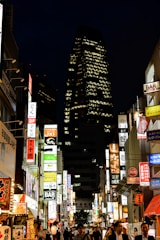 A bustling city street at night, illuminated by numerous colorful neon signs advertising various bars and businesses. The tall skyscraper in the background features a distinctive pattern of lights. People are walking through the lively street, capturing the energetic atmosphere of urban nightlife.