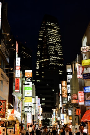 A bustling city street at night, illuminated by numerous colorful neon signs advertising various bars and businesses. The tall skyscraper in the background features a distinctive pattern of lights. People are walking through the lively street, capturing the energetic atmosphere of urban nightlife.