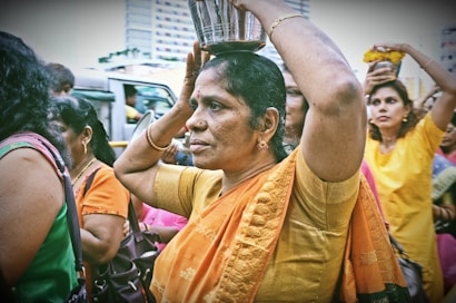 A group of women is participating in a cultural or religious procession. They are carrying metal vessels on their heads, adorned in colorful traditional attire. The background shows urban buildings and other participants.