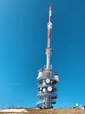 A tall, cylindrical broadcast tower stands against a clear blue sky. The structure is equipped with multiple antennas and satellite dishes, positioned on a grassy hill. There are several people nearby, highlighting the vastness of the tower.