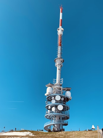 A tall, cylindrical broadcast tower stands against a clear blue sky. The structure is equipped with multiple antennas and satellite dishes, positioned on a grassy hill. There are several people nearby, highlighting the vastness of the tower.