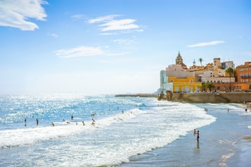 A picturesque coastal scene with people enjoying the ocean. The water sparkles under the bright sun, and waves gently hit the shoreline. In the background, colorful Mediterranean-style buildings with a prominent tower are positioned along a cliff, complemented by palm trees.