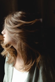 A person with light, wavy hair appears to be turning their head, causing the hair to flow dynamically in the air. The lighting is soft, highlighting the texture of the hair against a darker background.
