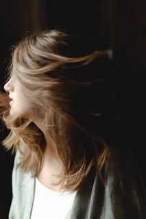 A person with light, wavy hair appears to be turning their head, causing the hair to flow dynamically in the air. The lighting is soft, highlighting the texture of the hair against a darker background.