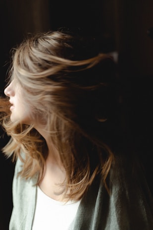 A person with light, wavy hair appears to be turning their head, causing the hair to flow dynamically in the air. The lighting is soft, highlighting the texture of the hair against a darker background.