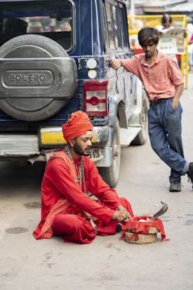 A man dressed in a vibrant red traditional outfit and turban sits on the ground near a woven basket with a snake. He is surrounded by necklaces and wears a serene expression. Another person, wearing a casual pink shirt and blue pants, leans against a dark blue SUV, observing the scene. The setting appears to be a street, with another person visible in the background reading a newspaper.
