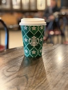 A green disposable coffee cup with white star patterns and a logo on a wooden table in a café setting. The background is softly blurred, suggesting a cozy atmosphere with additional tables and chairs.