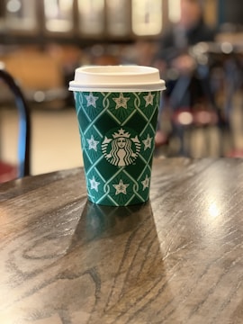 A green disposable coffee cup with white star patterns and a logo on a wooden table in a café setting. The background is softly blurred, suggesting a cozy atmosphere with additional tables and chairs.