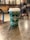 A green disposable coffee cup with white star patterns and a logo on a wooden table in a café setting. The background is softly blurred, suggesting a cozy atmosphere with additional tables and chairs.