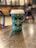 A green disposable coffee cup with white star patterns and a logo on a wooden table in a café setting. The background is softly blurred, suggesting a cozy atmosphere with additional tables and chairs.