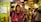 A bustling market scene with a young woman in a pink jacket holding a phone and looking around, surrounded by several other people walking and engaging with the market atmosphere. Bright, colorful lights and stalls create a lively backdrop.