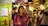 A bustling market scene with a young woman in a pink jacket holding a phone and looking around, surrounded by several other people walking and engaging with the market atmosphere. Bright, colorful lights and stalls create a lively backdrop.