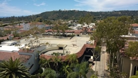 Aerial view of a suburban area with a mix of residential and commercial buildings. The scene includes red-tiled roofs, a parking area, and lush greenery with tall trees. A backdrop of hills and a partly cloudy sky can be seen in the distance.