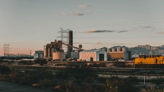 An industrial landscape with a large factory complex featuring multiple buildings, chimneys, and cooling towers. In the foreground, there is a yellow train on the railway tracks. The background showcases mountain ranges under a clear sky with scattered clouds. Power lines run across the scene, adding to the industrial setting.