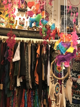 A vibrant and eclectic display featuring an assortment of colorful hanging decorations and dreamcatchers. The scene includes multicolored animal figures and feathers suspended from strings, as well as shelves lined with beckoning cat figurines. Below the decorations, a variety of patterned and colorful clothing is visible on hangers.