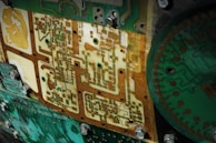 A close-up view of electronic circuit boards with detailed patterns and components. The intricate design includes various pathways, connections, and textured surfaces. Metallic bolts and nuts are visible, securing the pieces together, while the board exhibits a mix of green, brown, and cream colors.
