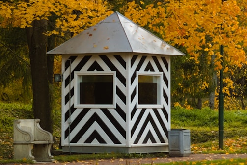 A small, striped black and white shelter with a pyramidal roof is set amidst a backdrop of vibrant yellow autumn leaves. There is a stone bench to the left and a small metal bin nearby, both resting on a paved surface. The lush greenery and trees surround the structure, contributing to an overall pastoral setting.